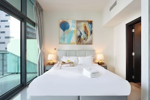 Une chambre d’hôtel avec un grand lit, deux tableaux au mur, une fenêtre avec des rideaux et une table de chevet avec une lampe.