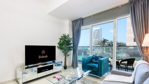 Una sala de estar moderna con un televisor de pantalla plana, una planta en maceta, un sofá color turquesa, una mesa de centro de vidrio y un balcón con vistas a un paisaje urbano.