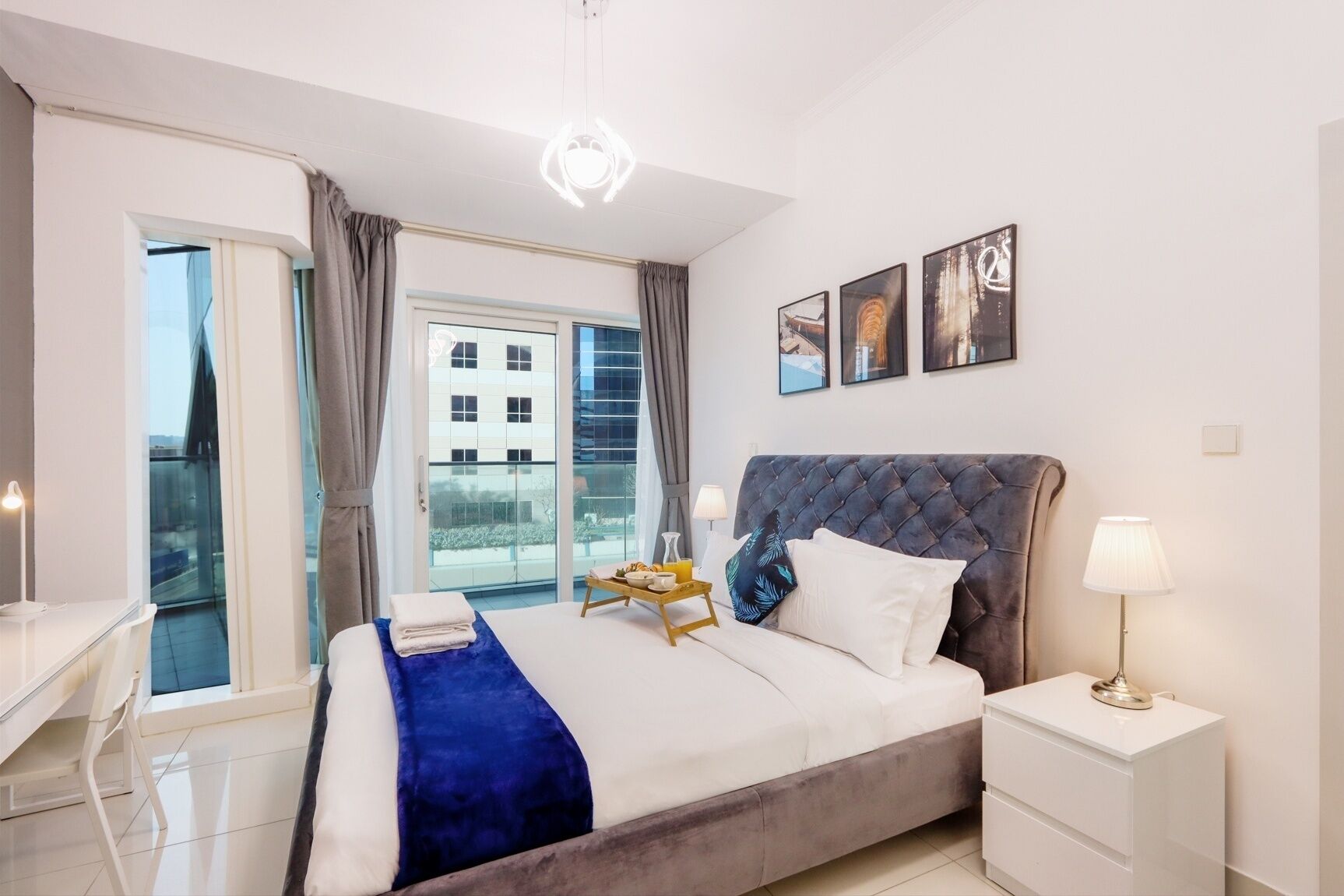 Une chambre moderne avec un grand lit, une table de chevet, un bureau et une vue sur un immeuble à travers une porte vitrée.