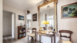 Une pièce comprenant un bureau en bois, deux chaises, une bibliothèque, un grand miroir et un tableau.