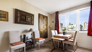 Un salon moderne avec un coin repas, doté d’un parquet, de meubles blancs et de grandes fenêtres offrant une vue sur la ville.