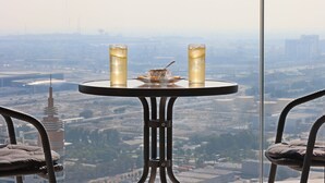A table with two glasses of a beverage, a glass of water, and a small bowl with a spoon, set against a cityscape view from a high vantage point.