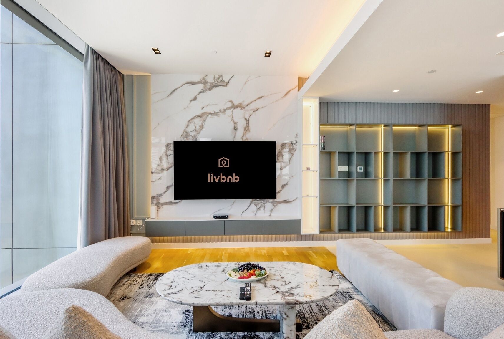A modern living room with a marble-patterned wall, a flat-screen TV, a coffee table with fruits, and a built-in shelving unit.