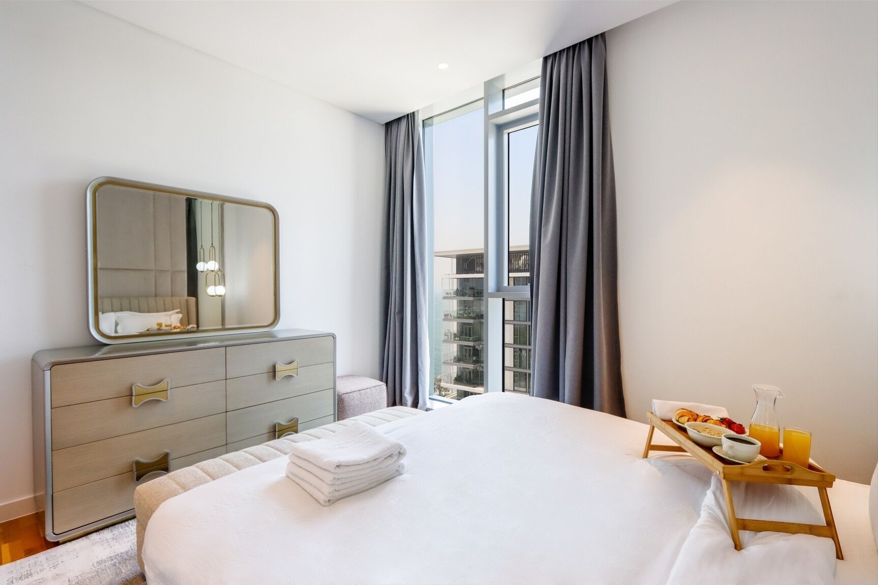 Habitación de hotel moderna con una cama grande, un aparador con cajones, un espejo, una mesita de noche con una bandeja de desayuno, y vistas a la ciudad a través de amplias ventanas.