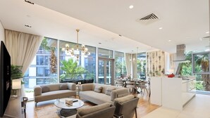 Una sala moderna con un sofá seccional grande, una mesa de centro y un televisor. Hay un comedor con mesa y sillas, y una cocina con mostrador y lavamanos. La habitación tiene ventanas grandes con vistas a palmeras y edificios.