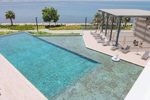 A swimming pool with clear water, a lounge area with sunbeds and umbrellas, and a view of the sea.