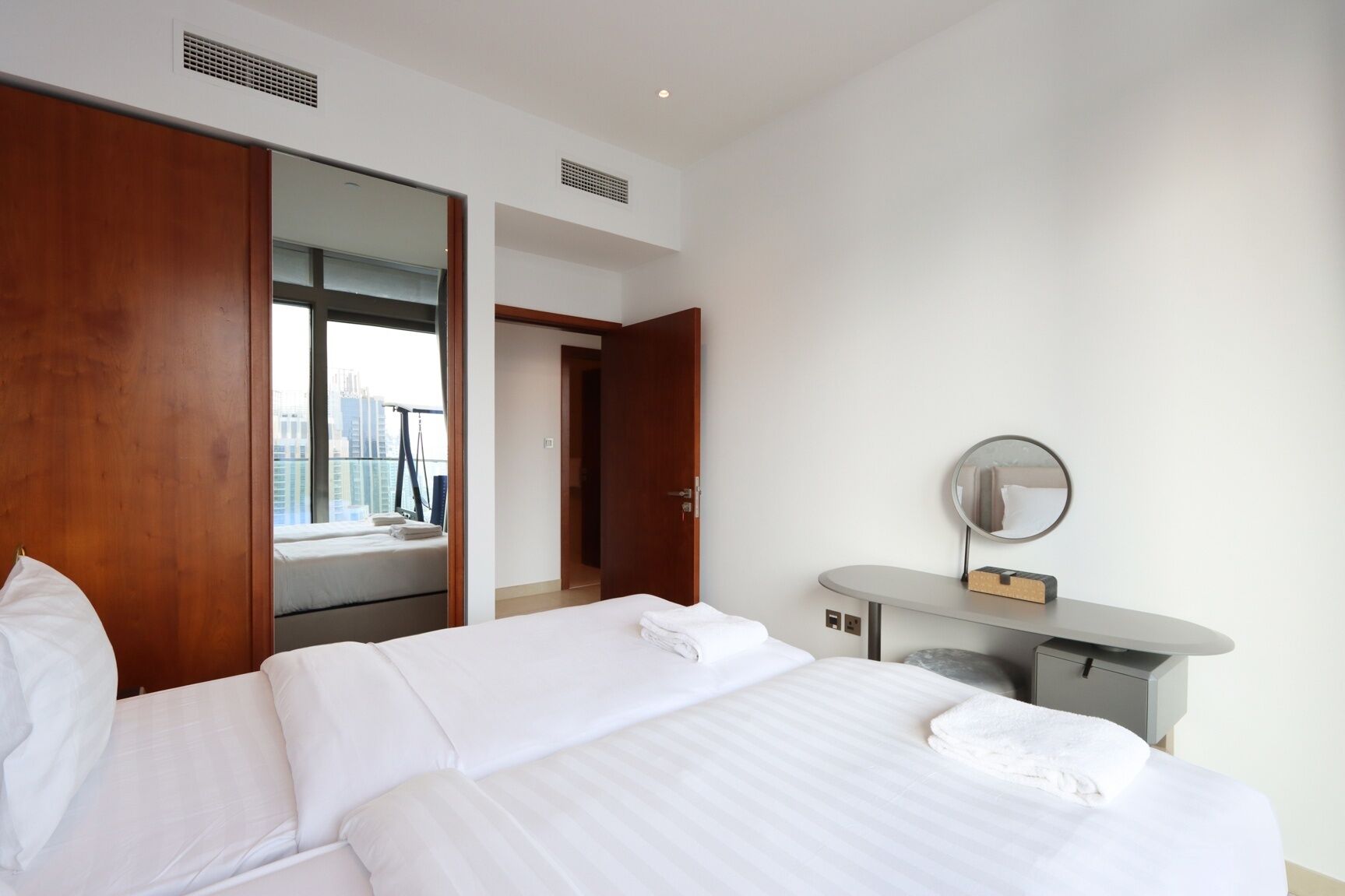 Ein Hotelzimmer mit zwei Betten, einem Schreibtisch mit Spiegel und Blick auf die Stadt durch ein großes Fenster.