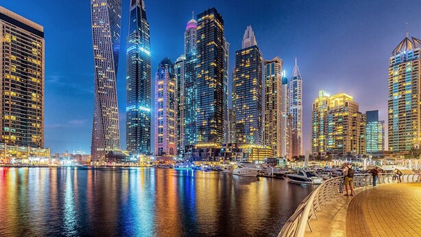 Stadsgezicht aan het water met moderne wolkenkrabbers, een jachthaven met boten en een boulevard.