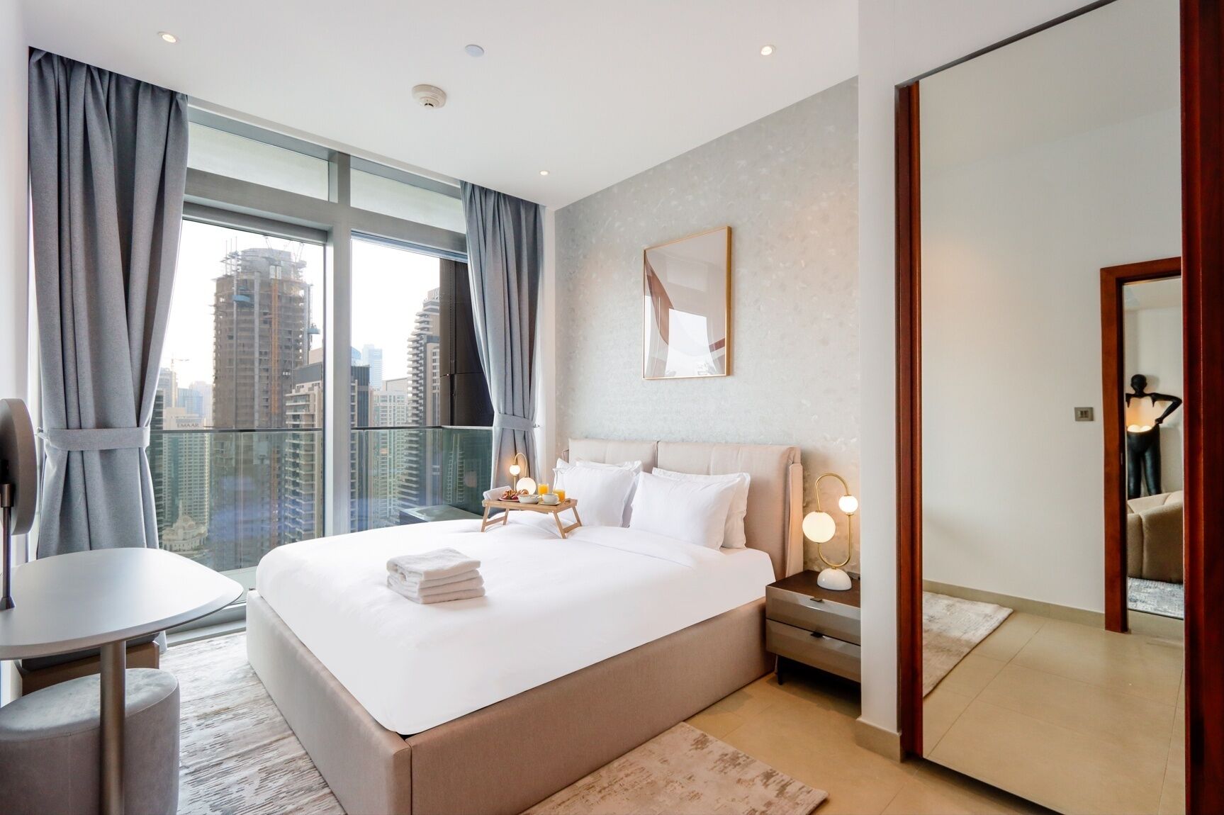 Een moderne hotelkamer met een groot bed, een nachtkastje met lamp, een klein tafeltje met handdoeken en uitzicht op de stad door grote ramen.