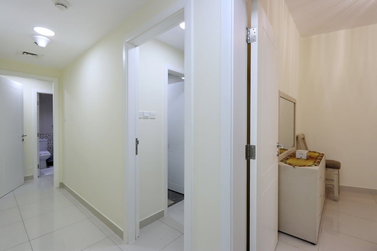 Un pasillo con paredes blancas y puertas que dan a un baño y una pequeña zona de estar con un espejo y un taburete.