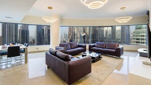 Sala de estar moderna com sofás de couro, mesa de centro de vidro e iluminação pendente. Janelas amplas oferecem vista para a cidade.