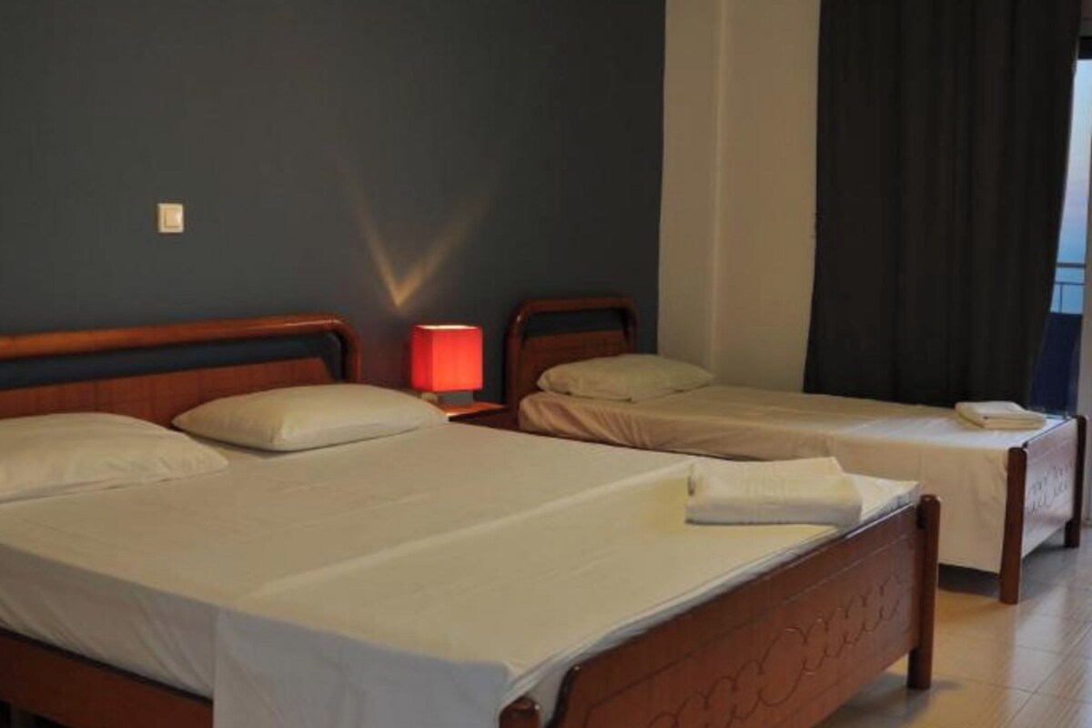 A hotel room with three wooden beds, each with white linens and pillows. There is a nightstand with a red lamp between the beds. The room has a dark blue wall and a window with dark curtains.