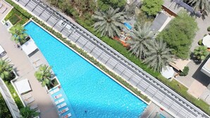 An aerial view of a rectangular swimming pool with a surrounding walkway, palm trees, and a building with a flat roof.