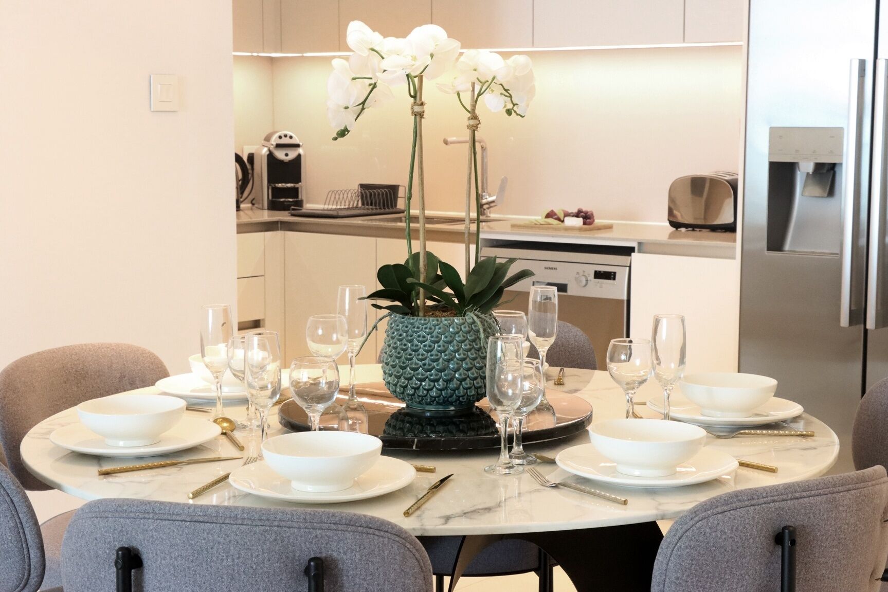 A modern dining area with a round table set for a meal, featuring white plates, glasses, and cutlery. A decorative pineapple plant is placed in the center of the table.