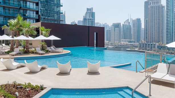 Una piscina infinita en la azotea con sillones de descanso y sombrillas, rodeada de edificios altos.