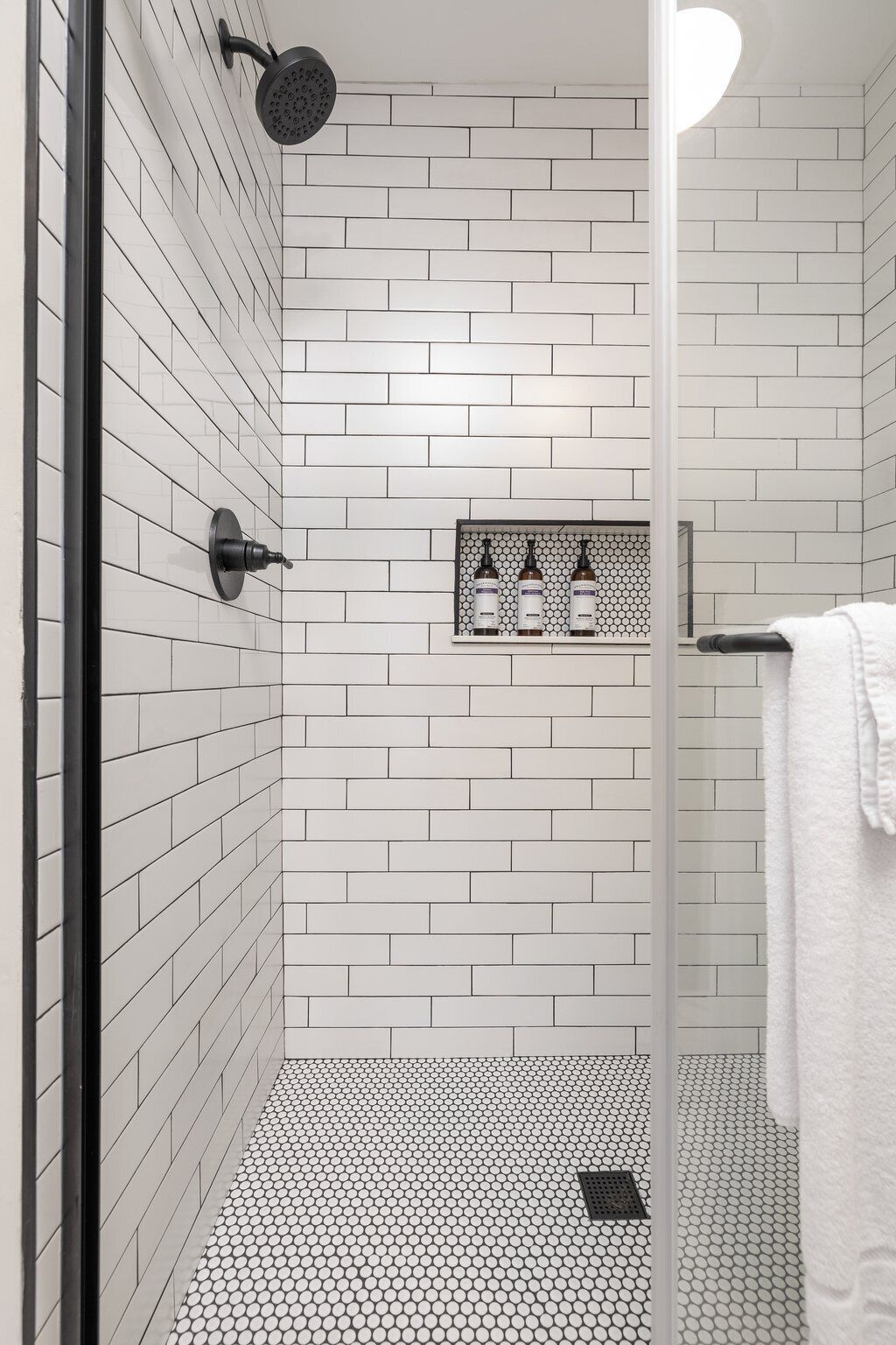 Un baño moderno con azulejos blancos tipo metro, una ducha con rociador negro y un estante con tres botellas.