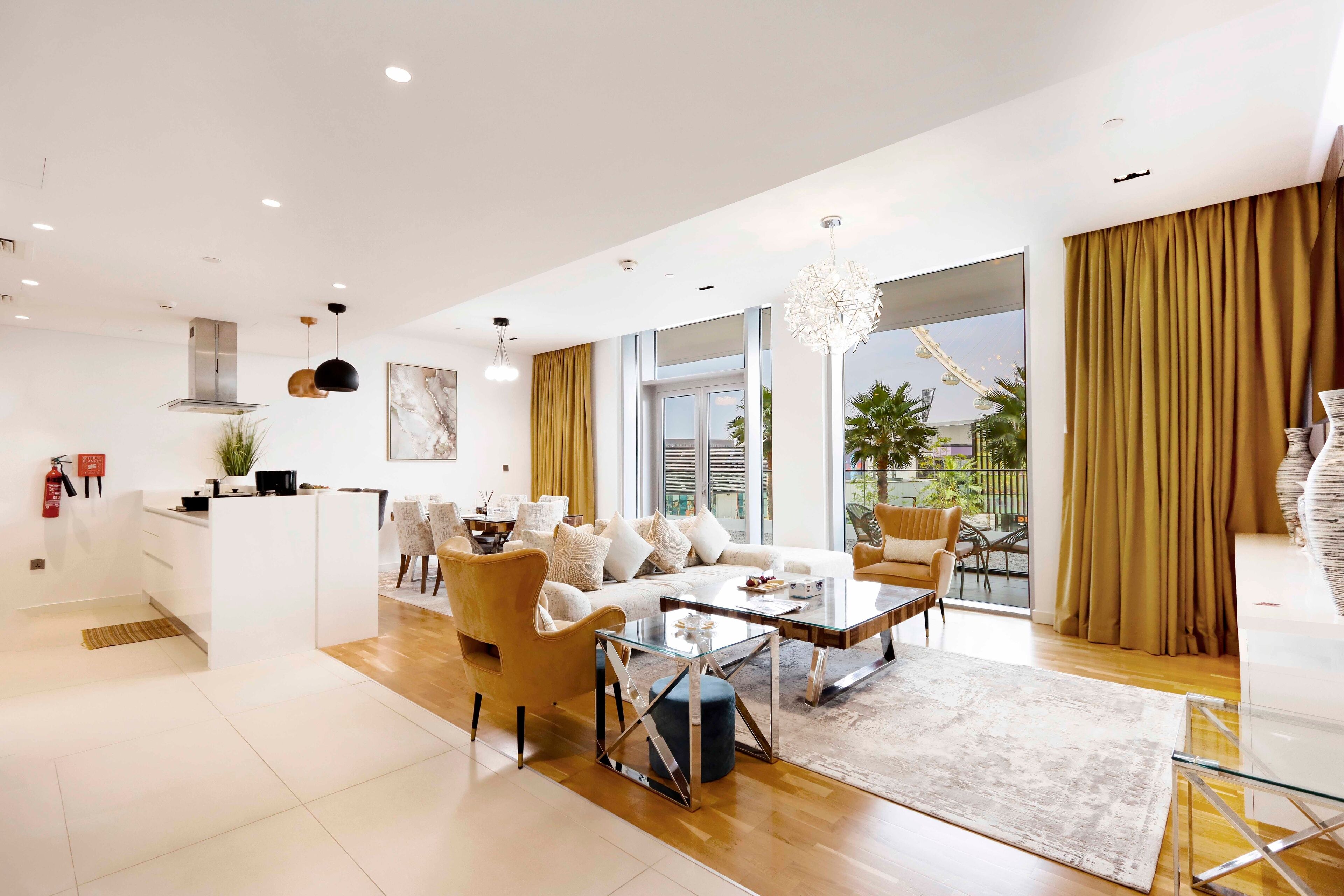 A modern living room with a sofa, coffee table, and chairs, a kitchen area, and large windows with curtains.