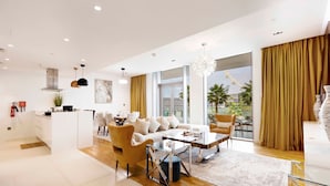 A modern living room with a sofa, coffee table, and chairs, a kitchen area, and large windows with curtains.