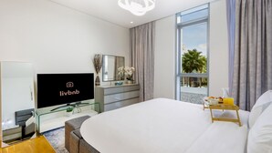 Ein modernes Hotelzimmer mit einem großen Bett, einem Flachbildfernseher, einem Schminktisch, einem Spiegel und einem Balkon mit Blick auf Palmen.