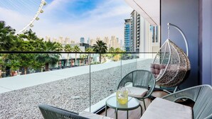 A balcony with wicker chairs, a hanging chair, and a table with a pitcher of drink. A cityscape with high-rise buildings is visible in the background.