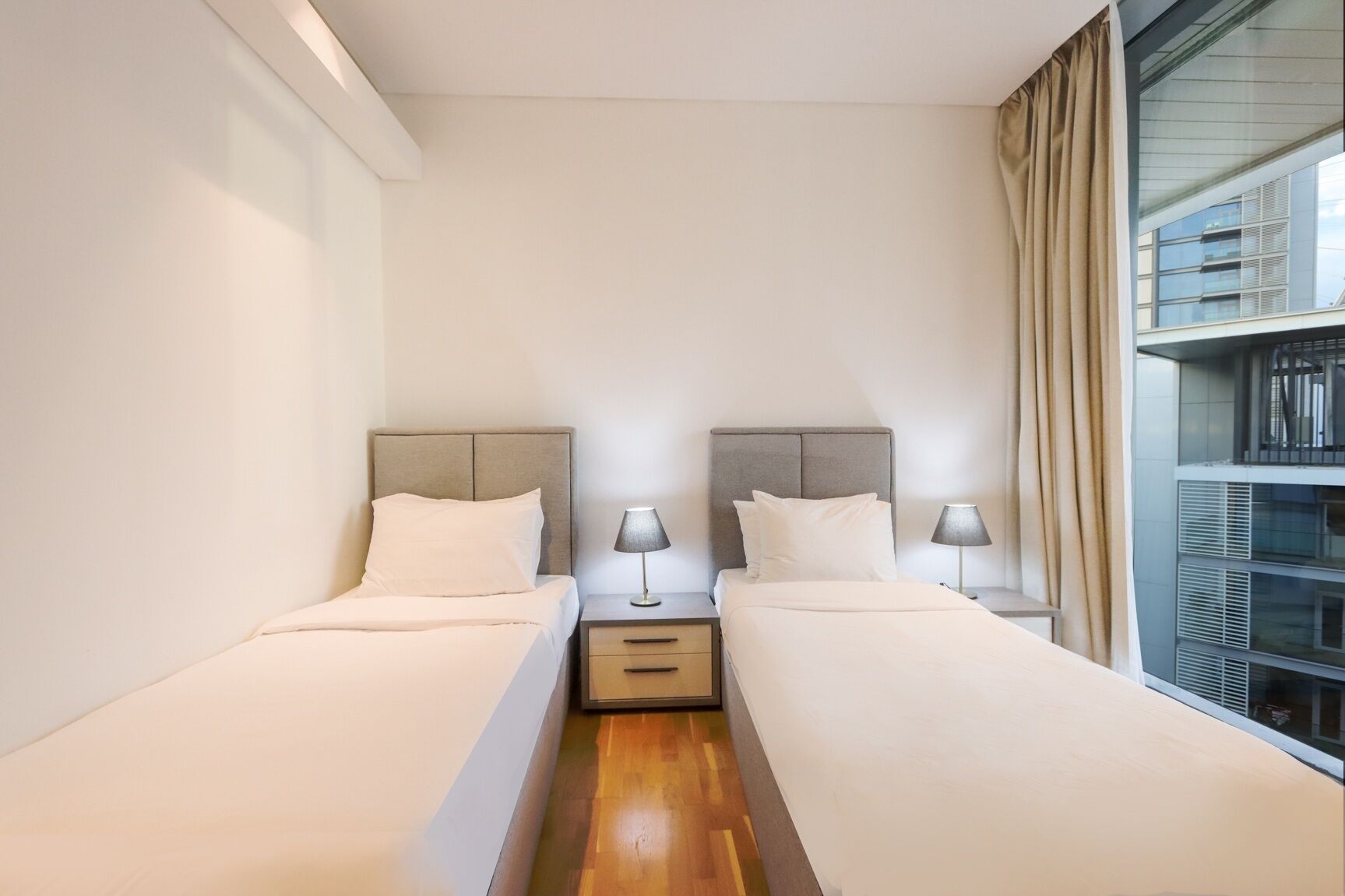 Una camera d'albergo con due letti singoli, un comodino con una lampada e un'ampia finestra con tende.