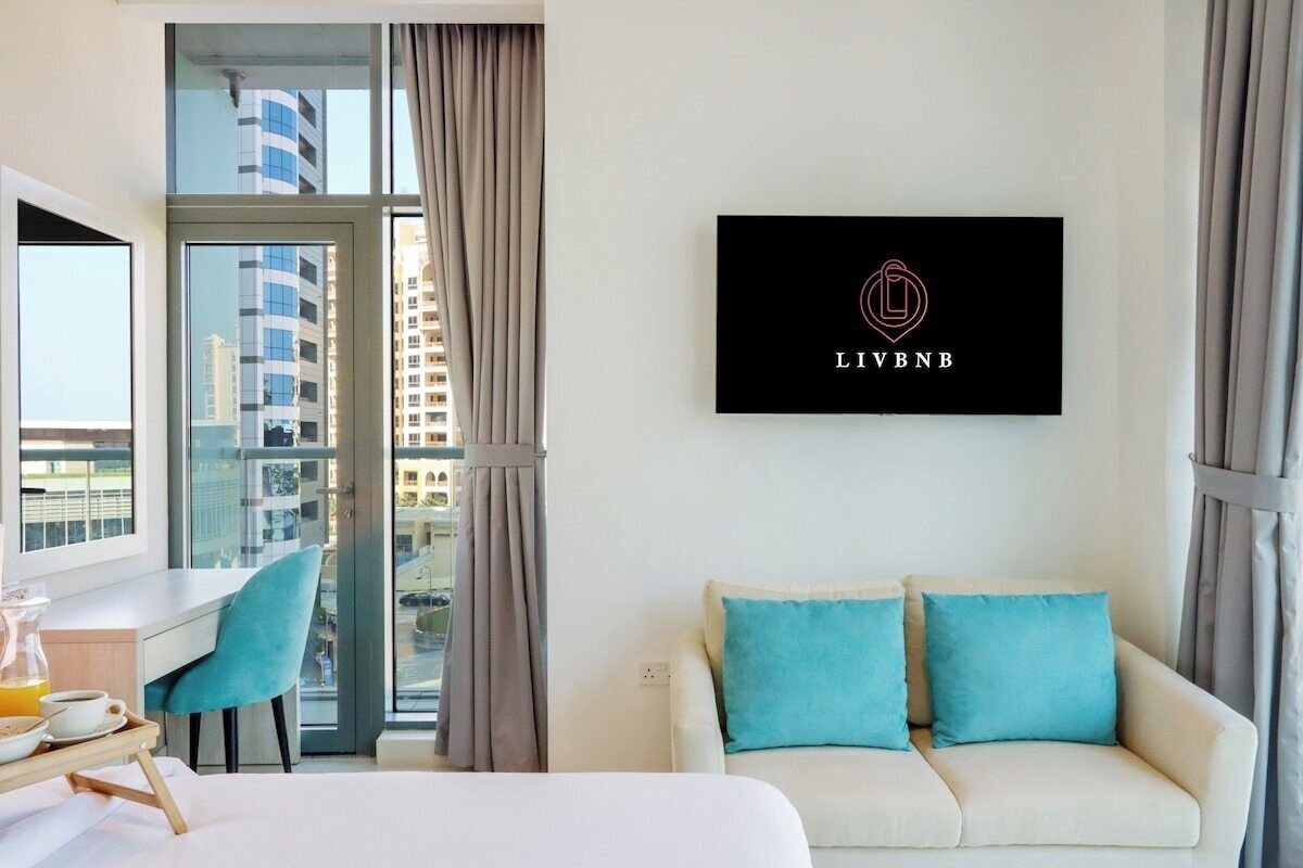 Ett hotellrum med en säng, ett skrivbord, en stol, en soffa med kuddar, ett fönster med gardiner och en väggmonterad skylt med en logotyp.