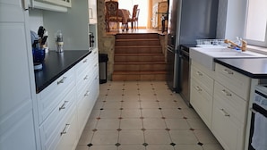 Une cuisine avec des meubles blancs, un plan de travail noir et un sol carrelé. Un escalier mène à un coin repas équipé de chaises et d’une table.