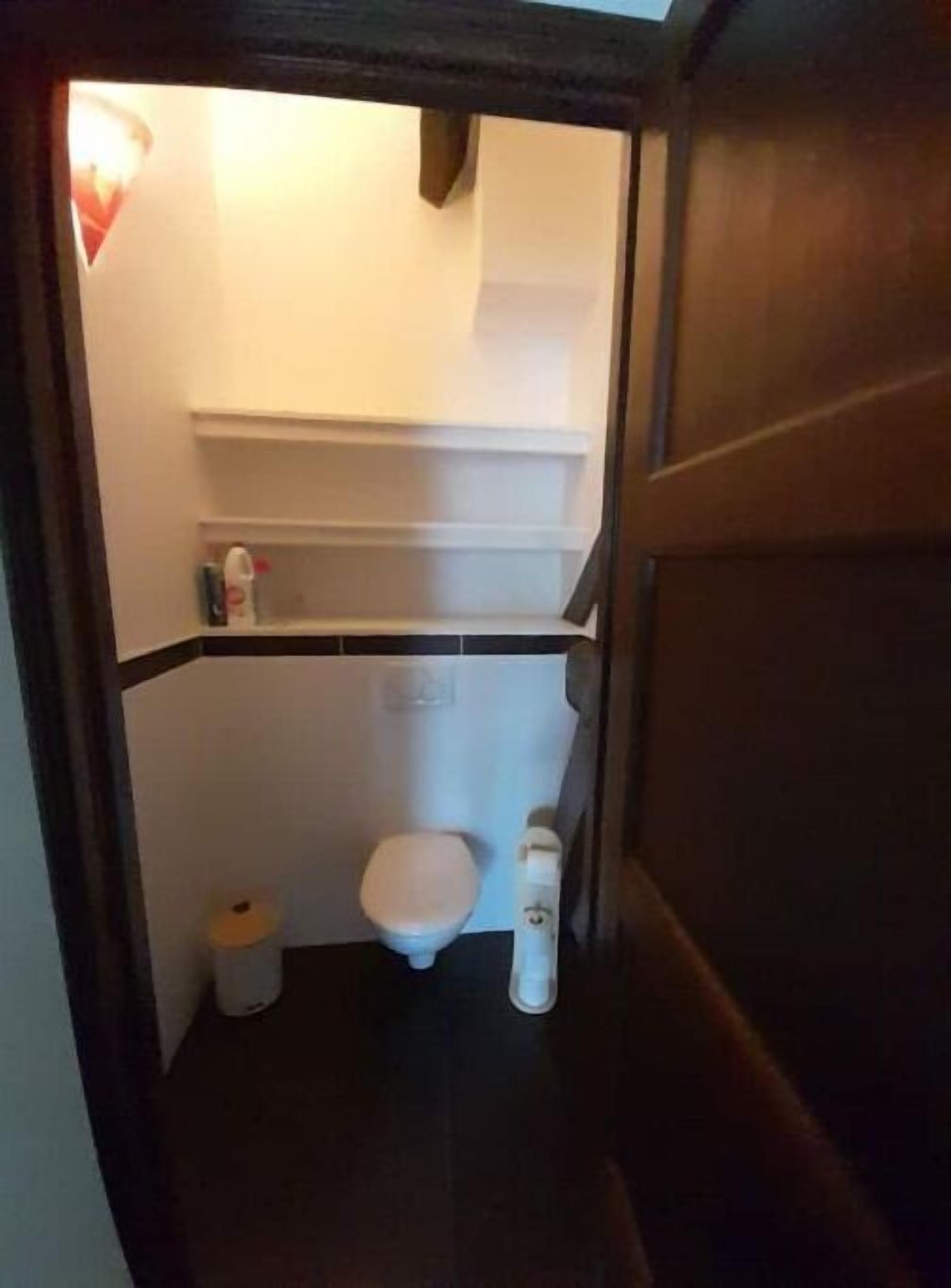 Um banheiro pequeno e estreito com vaso sanitário, uma prateleira acima do vaso e uma lixeira.