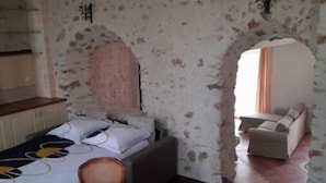 Une pièce avec un mur en pierre, un lit recouvert d’une couvre-lit à motifs, une table en verre avec des chaises et un canapé avec des coussins.