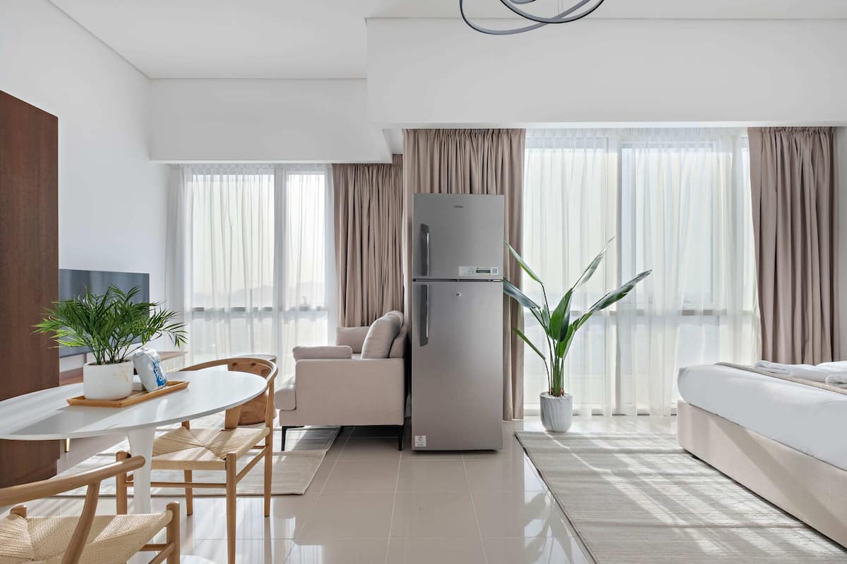 Una sala de estar moderna con un sofá, una mesa de comedor con sillas, un refrigerador y ventanas grandes con cortinas.