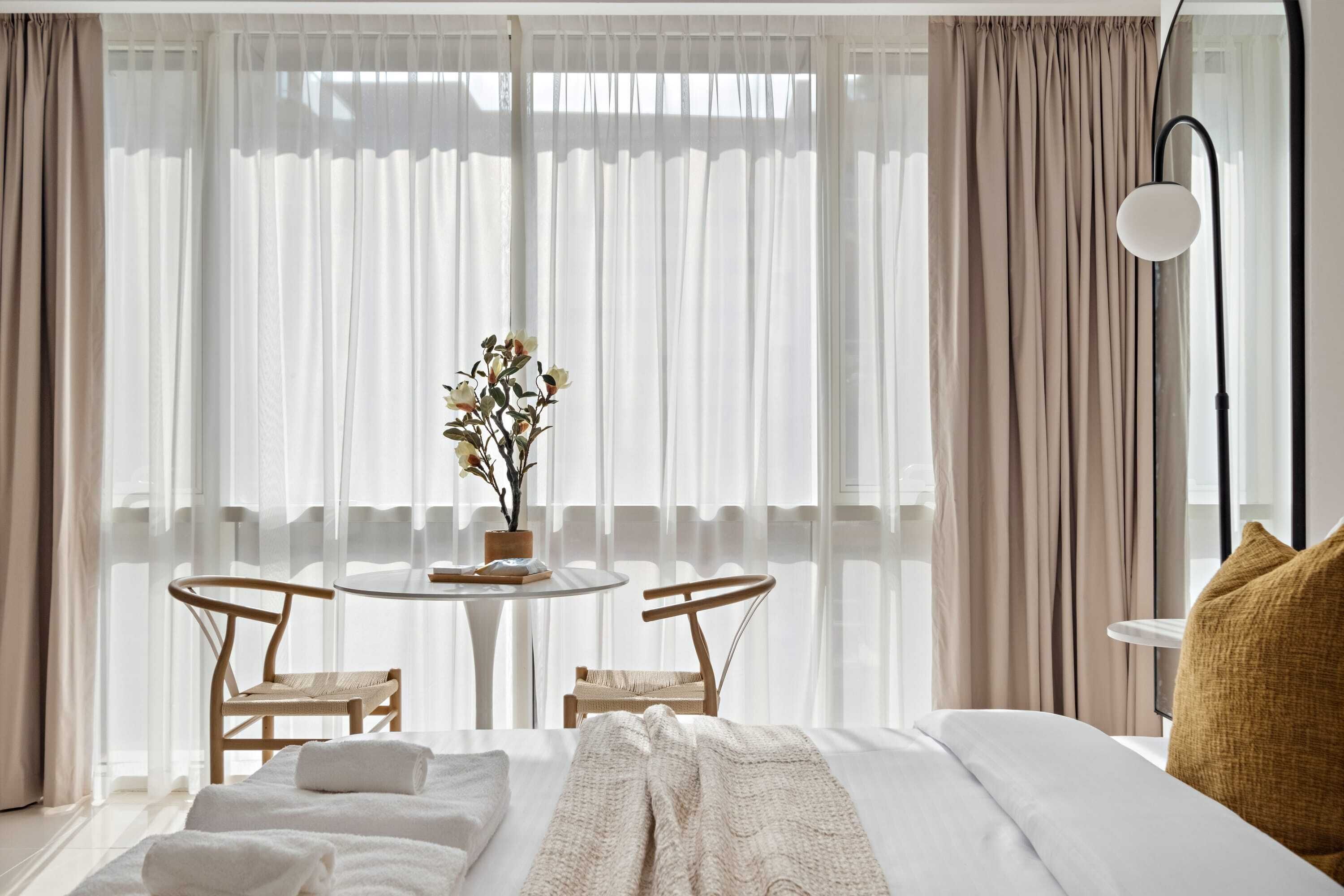 Ein Hotelzimmer mit einem Bett, zwei Stühlen, einem Tisch mit Blumenvase und leicht transparenten Vorhängen.
