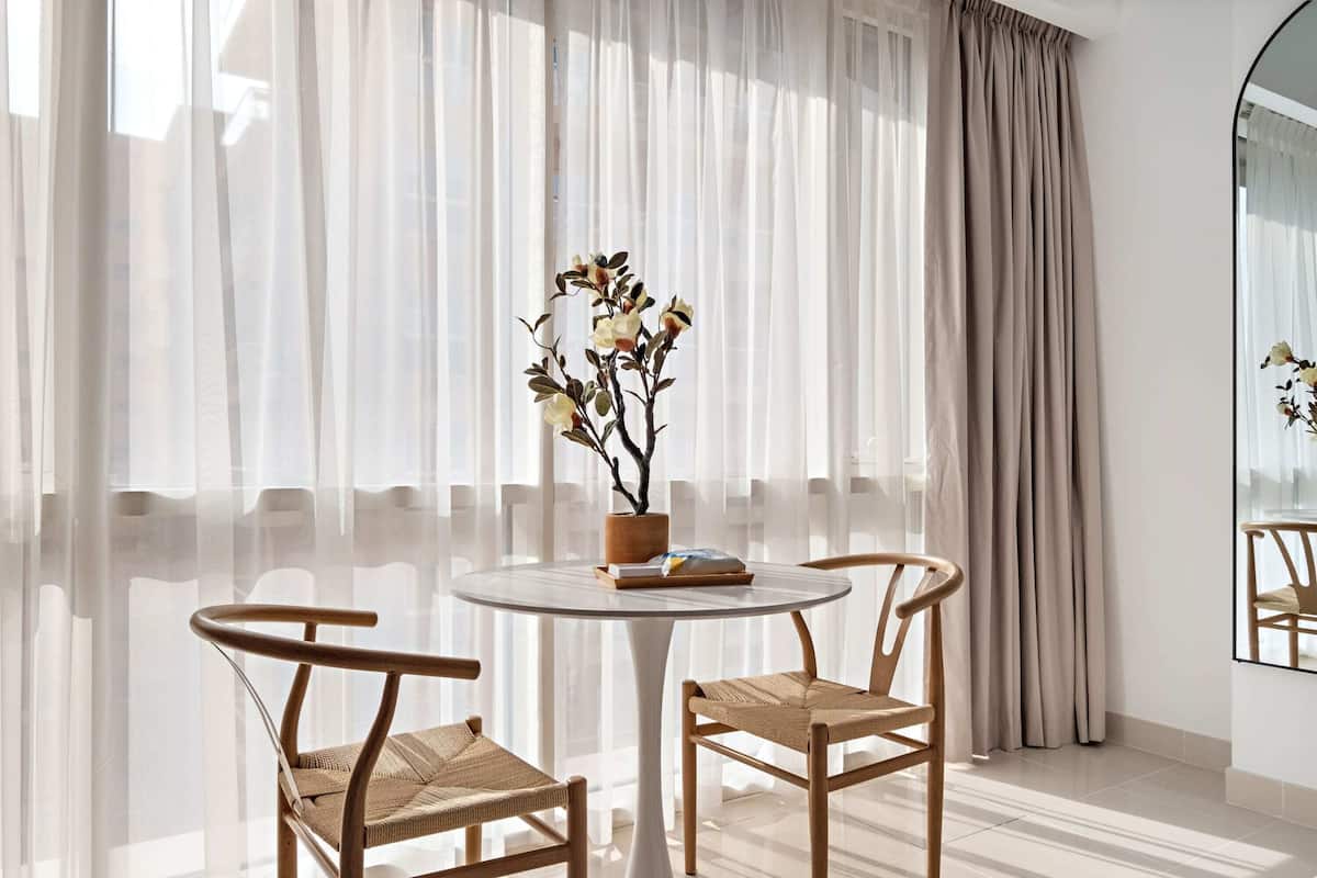 Una habitación con una mesa redonda, dos sillas de mimbre, una planta en maceta y cortinas de encaje.