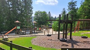 Une aire de jeux dotée de diverses installations, notamment des balançoires et un toboggan, entourée d’arbres et d’une clôture en bois.