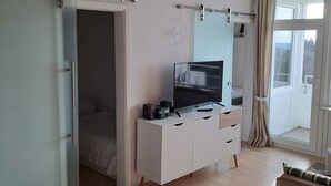 Une chambre d’hôtel avec un lit, un téléviseur posé sur un meuble blanc, une petite table avec une chaise et un balcon avec des rideaux.