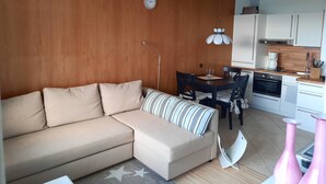 Un salon moderne avec un canapé beige, une table à manger avec des chaises, un coin cuisine doté de meubles blancs et un mur d’accent en bois.