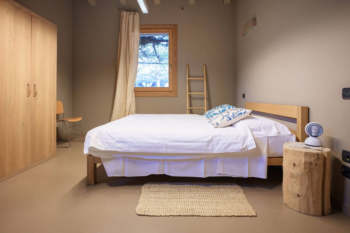 A bedroom with a bed, a wooden wardrobe, a window with curtains, a wooden ladder, and a bedside table with a lamp.