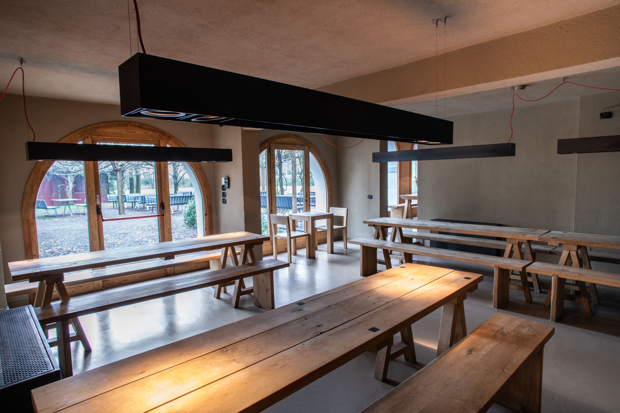 A spacious indoor area with wooden benches and tables, large windows, and a modern ceiling light fixture.