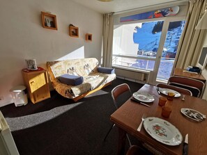 A cozy room with a sofa bed, a dining table with chairs, a small cabinet, and a window with a view of a snowy landscape.
