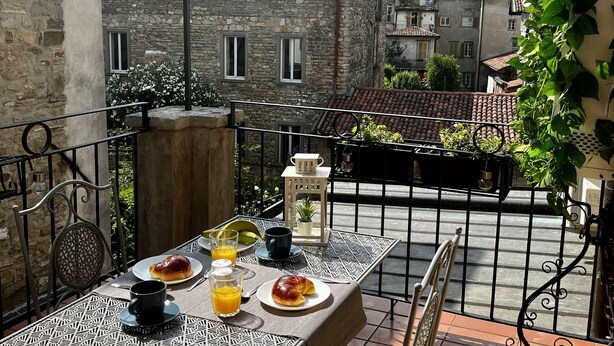 En balkong med ett bord dukat för frukost, med bland annat croissanter och apelsinjuice, med utsikt över en historisk stad med byggnader i sten och en kyrka.