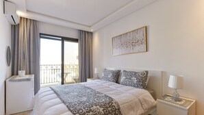 Ein Hotelzimmer mit einem großen Bett, einem Nachttisch mit Lampe, einem kleinen Beistelltisch, einem Fenster mit Vorhängen und einem Bild an der Wand.
