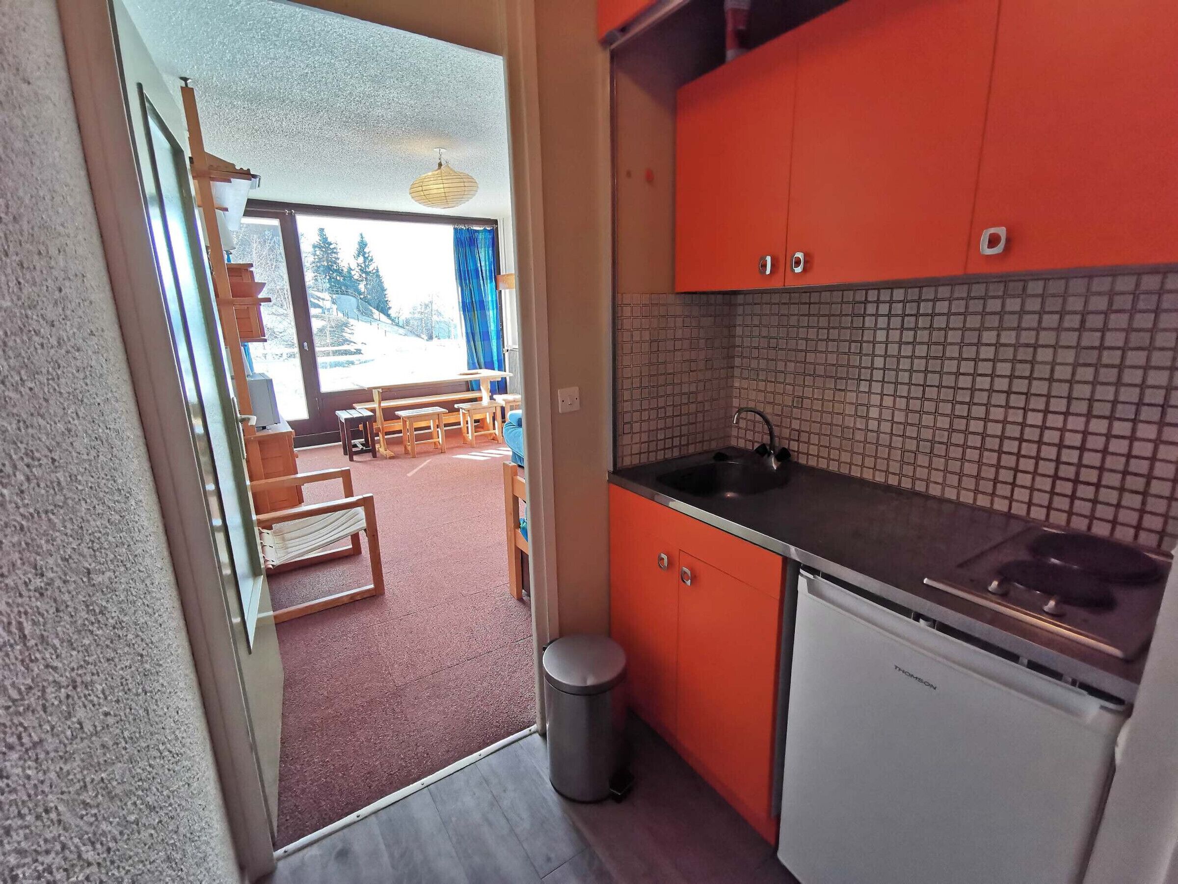 A compact kitchen with orange cabinets, a tiled backsplash, and a view of a dining area through an open door.