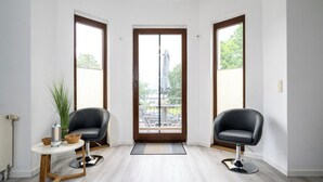 Una habitación con piso de madera, dos sillas negras, una mesita con un cuenco y un calentador, y una puerta que da a un espacio exterior con un banco y una sombrilla.