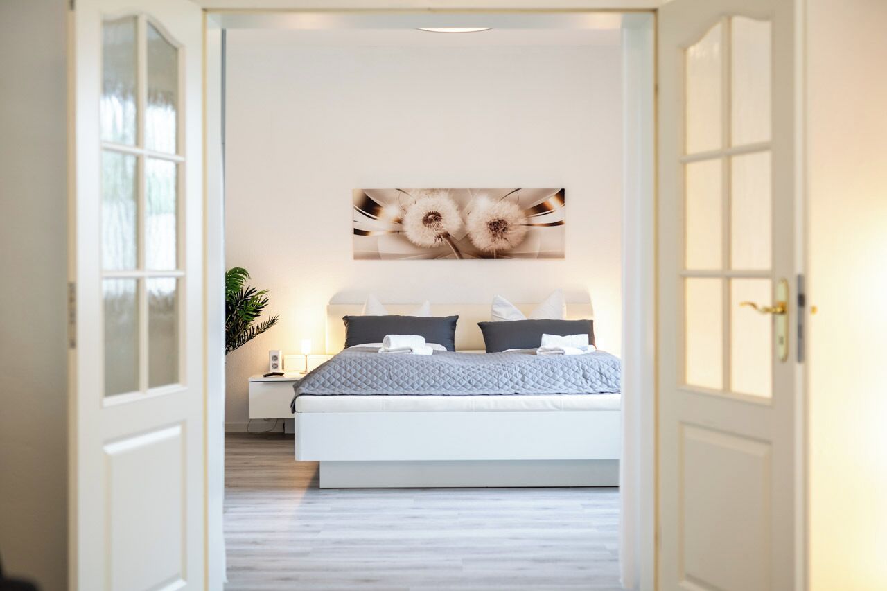 Ein Schlafzimmer mit Bett, Nachttisch, Blumentopf und einem an der Wand hängenden Kunstwerk.