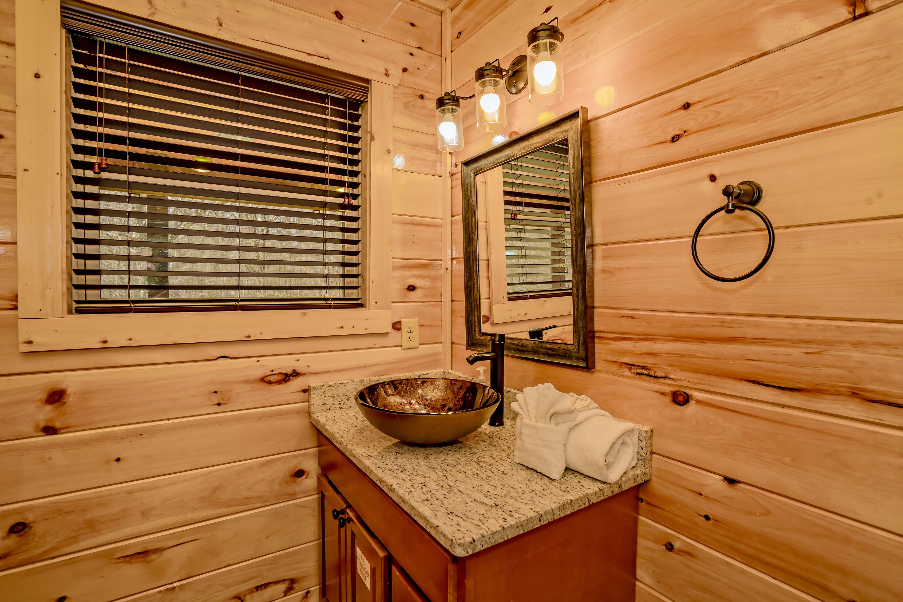 Un baño de cabaña de madera con encimera de granito, un espejo grande y una ventana con persianas.