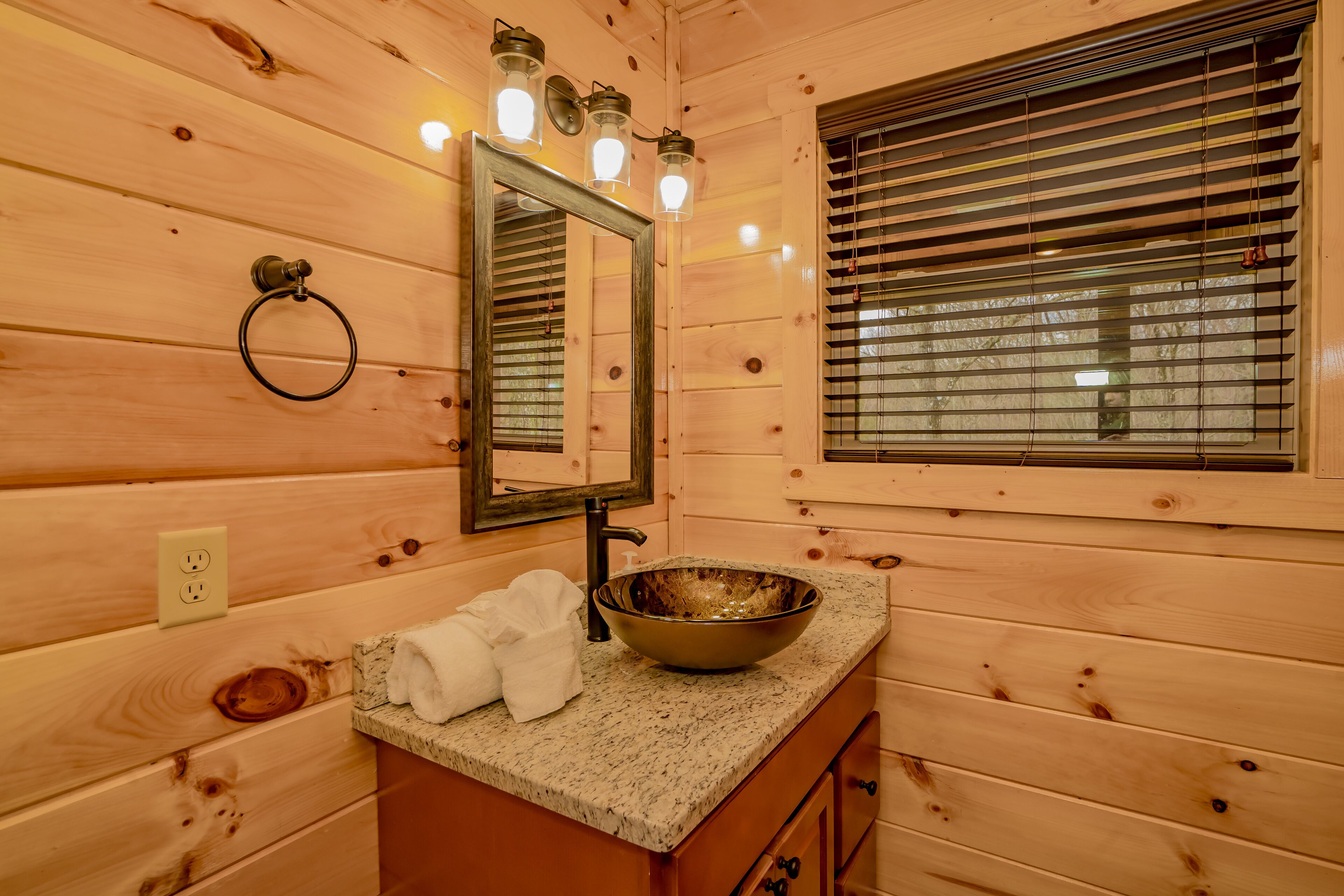 Une salle de bain rustique, avec des murs en bois, un comptoir en granit, un lavabo, un miroir et une fenêtre équipée de stores.