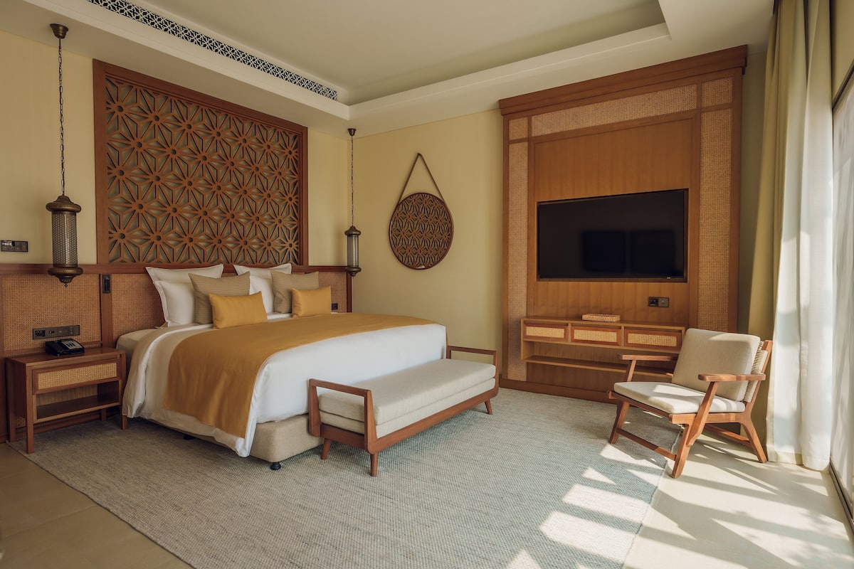 Ein modernes Hotelzimmer mit einem großen Bett, einem Fernseher, einem Holzstuhl und einer dekorativen Wandverkleidung.