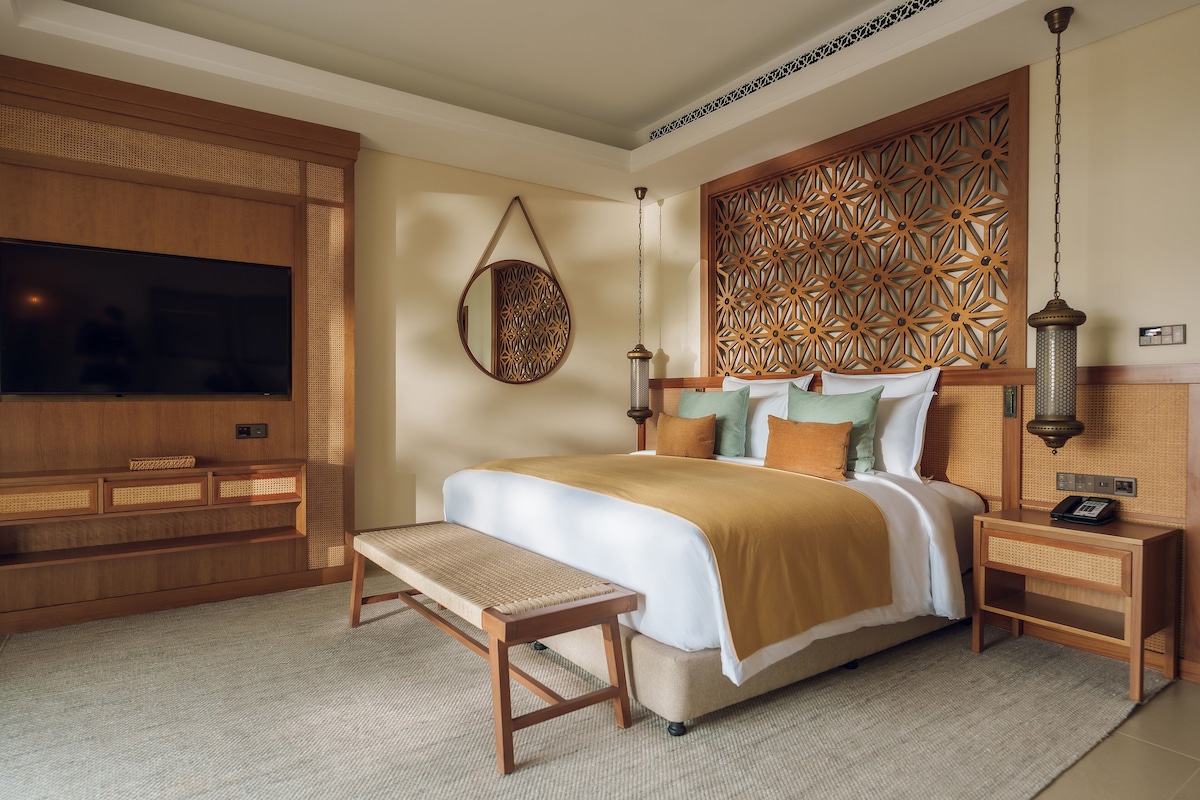 A bedroom with a large bed, a TV, a hanging mirror, and a decorative wall panel.