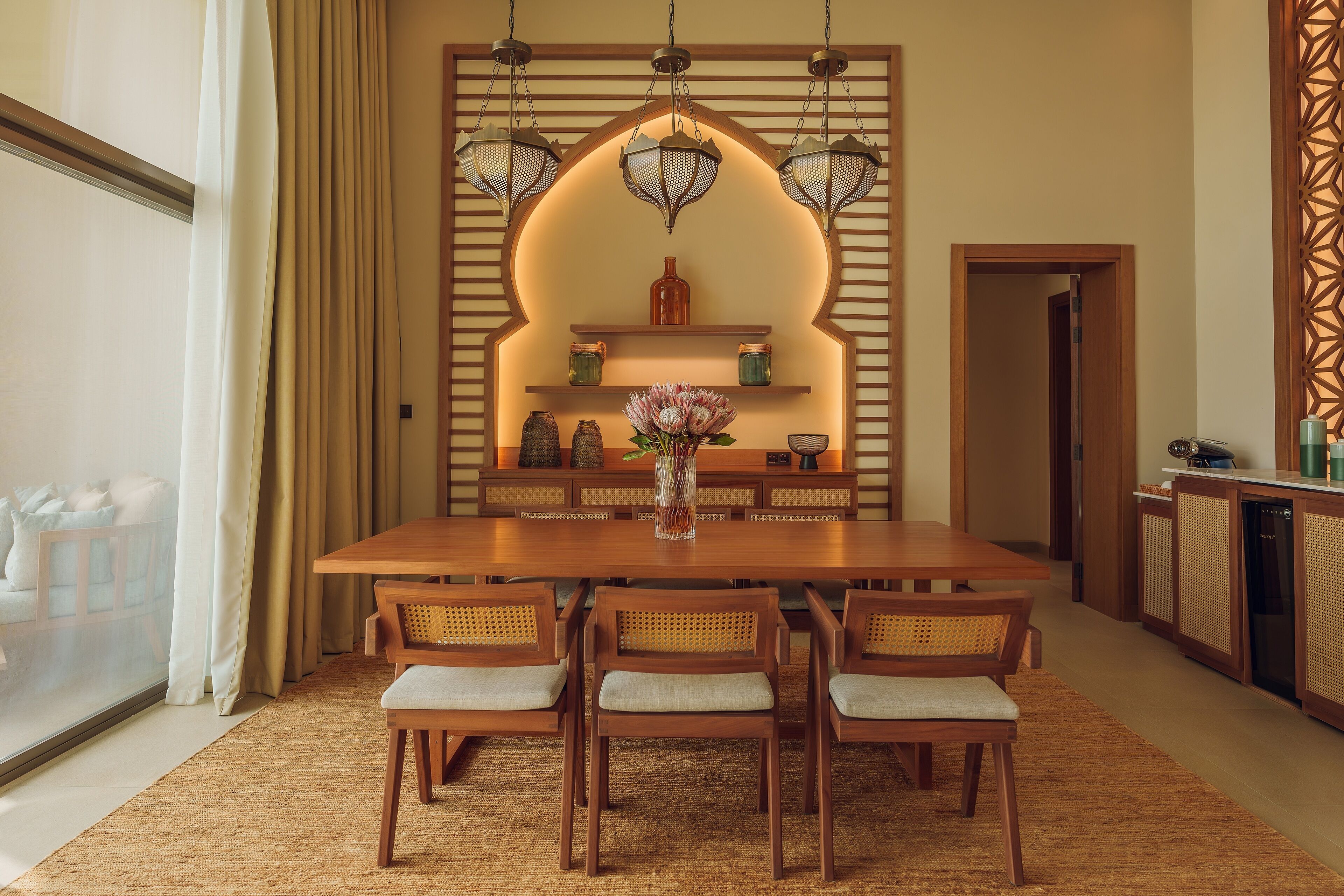 Un comedor con una mesa de madera, cuatro sillas, un jarrón con flores y una pared decorativa con estantes y luces colgantes.