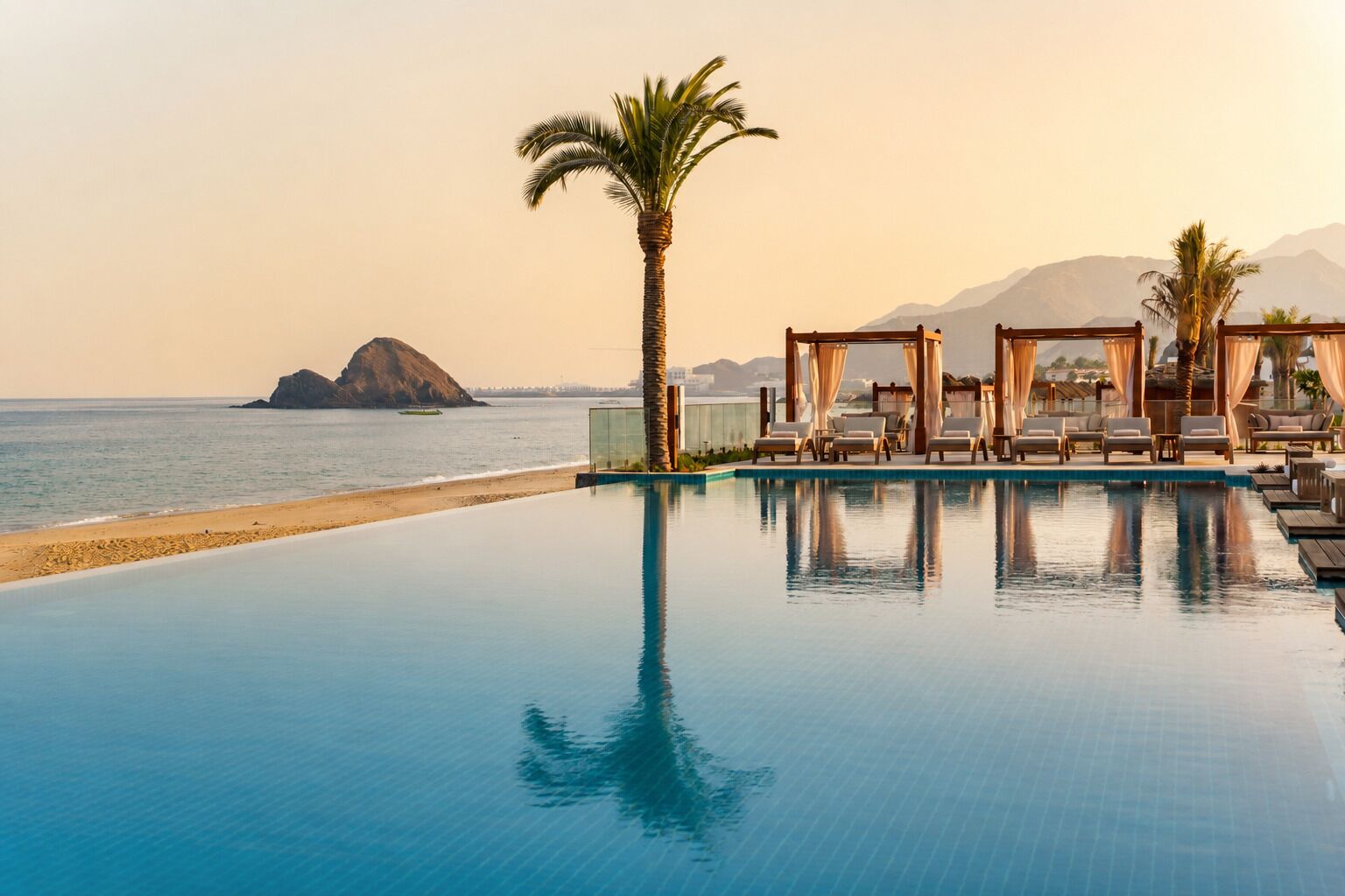 Un espace aménagé au bord de la piscine, avec des chaises longues et un palmier, offrant une vue sur la plage et les montagnes.