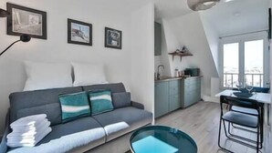 A modern apartment with a grey sofa, teal cushions, a glass coffee table, and a kitchen area visible through an open door.